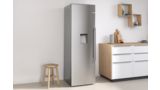 Silberfarbener freistehender Bosch Kühlschrank zwischen einem kleinen Stuhl links und einem Sideboard rechts.