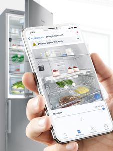 Téléphone avec l'application Home Connect montrant une alerte « Veuillez fermer la porte » et une porte de réfrigérateur ouverte en arrière-plan.