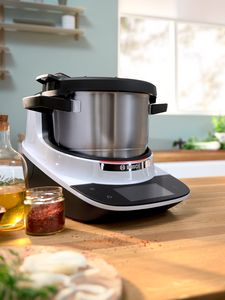 The Cookit cook processor stands on a wooden worktop, next to it oil and spices and a board.