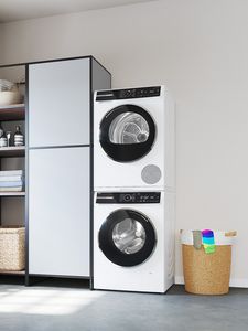 In einem hellen Raum steht ein Wäschetrockner auf einer Waschmaschine mit einem Schrank daneben.