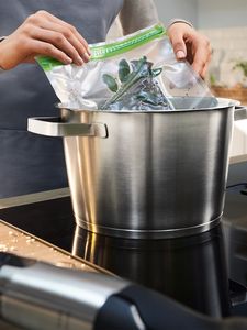 Mãos a segurar um saco de legumes selado a vácuo sobre uma panela, preparando-se para os cozinhar a vácuo.