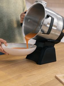 Le bol Cookit est fixé au nouveau socle verseur Cookit et incliné sur le côté gauche. Une personne tient le bol d'une main tout en vidant la soupe de carottes du bol dans une soupière de l'autre main.