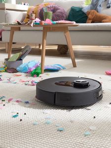 robot sprzatajacy na dywanie, w stle konfetti, stol i zabawki