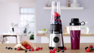 Mini blender Bosch VitaPower Series 2 cu fructe roșii și o sticlă To-Go plină cu smoothie pe un raft de bucătărie.