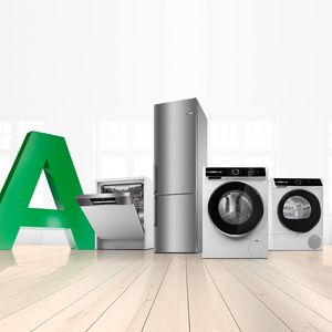 Eine Spülmaschine, ein Kühlschrank, eine Waschaschine und ein Trockner stehen in einem hellen Raum neben einem großen grünen A, stellvertretend für Energieeffizienzklasse A.