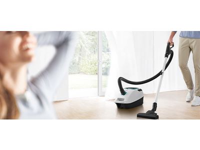 Two Bosch bagged cylinder vacuums stand on a cream rug in a light, bright living room.