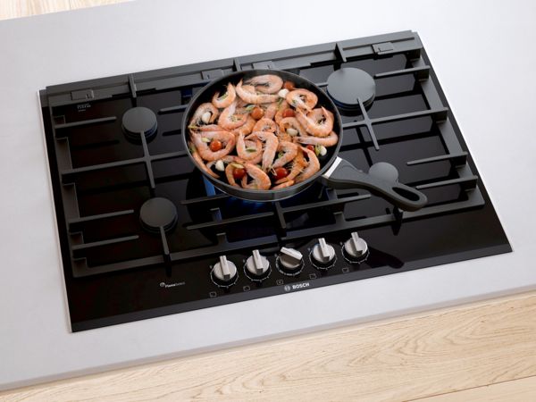Bosch 75 cm-es fekete edzett üveg gázfőzőlap egy teli serpenyő főzésben levő garnélarákkal és zöldséggel a közepén.