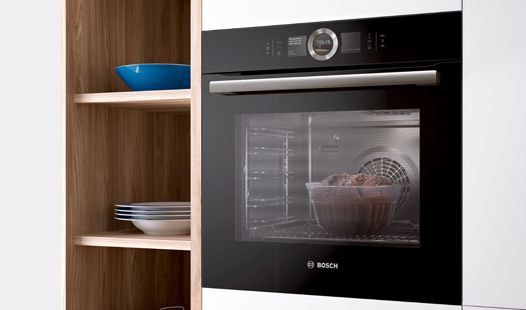 תנור של Bosch ועוגה הנאפית בתוכו ומדפים עם כלים המייצגים את מכשירי הבישול והמכשירים הביתיים של Bosch.