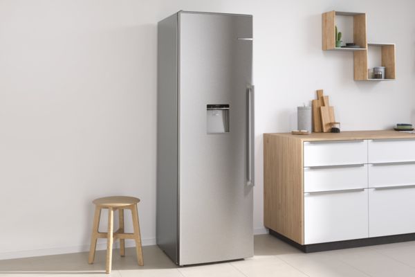 Серебристый отдельностоящий холодильник Bosch между небольшим табуретом слева и буфетом справа.