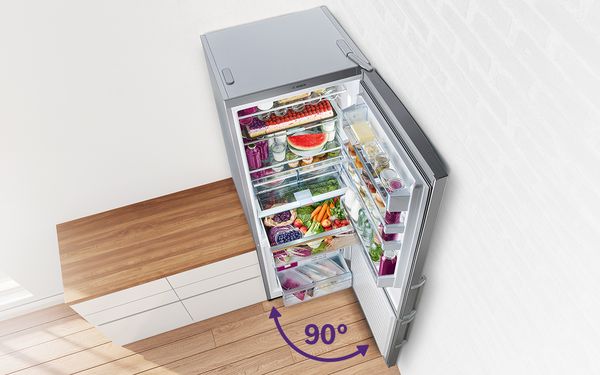 Открытый большой отдельностоящий холодильник с морозильной камерой в углу комнаты. Дверца холодильника, шириной 70 см, открыта на 90 градусов, в холодильнике лежат продукты питания. 