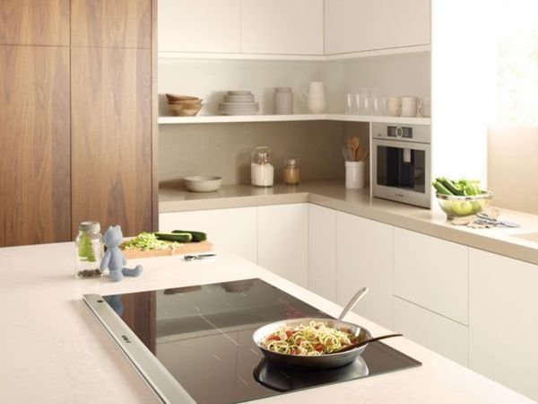 Minimalistinė baltos spalvos virtuvė su spintelėmis be rankenėlių bei įmontuota indukcine kaitlente ant kurios padėta maža keptuvė su makaronų patiekalu