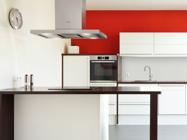 Bright red wall above the upper cabinets of a modern loft-style compact kitchen with white cabinets and built-in stainless-steel appliances.