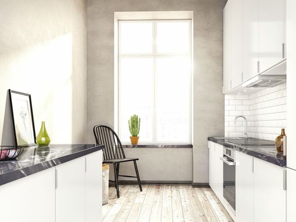 Izgaismota kambīzes tipa virtuve ar atklātām betona sienām, spīdīgiem baltiem skapīšiem, melnām marmora letēm un lielu logu telpas galā