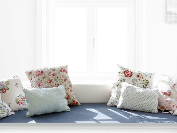 Cozy bay window with an upholstered blue window seat and white and flowered pillows in a small galley kitchen.