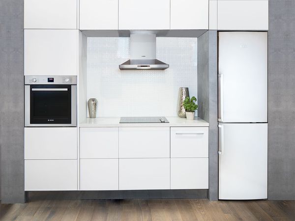 Small white fridge freezer installed flush with a concrete wall in a tiny, modern kitchen with glossy white cabinets