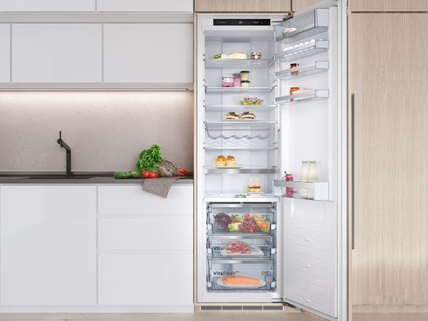 Réfrigérateur encastré avec à côté des ingrédients pour une salade sur un plan de travail et une crédence rosée   