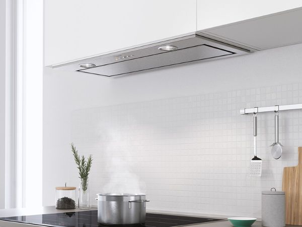 Steam rising into a Bosch canopy hood above a counter with spices and utensils hanging on a wall rack in front of a small tile backsplash