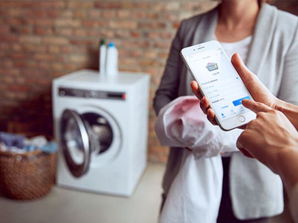 Smartphone cu aplicația Home Connect folosit pentru controlul unei mașini de spălat rufe Bosch pe fundal