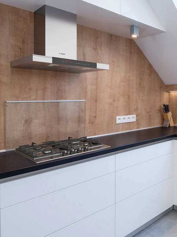 L-vormige keuken met witte kasten en een zwart werkblad onder een schuin dak met lichtkoepels. Elegante houten achterwand met verticale nerf