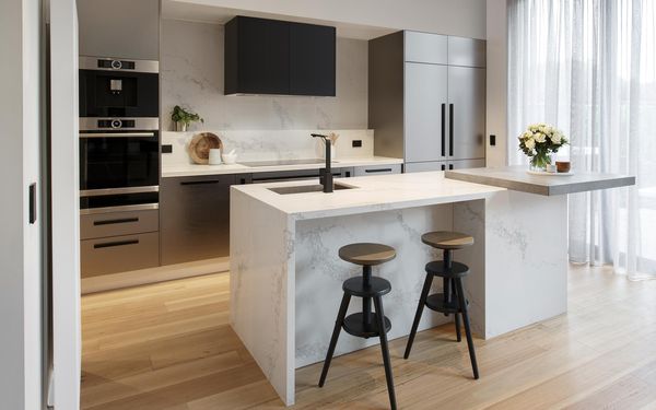 Bucătărie cu insulă din marmură și panou de protecție la stropire albe și dulapuri gri, cu suprafață reflectorizantă. Bucătăria prezintă un cuptor, plită și hotă.