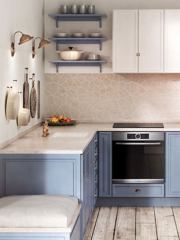 Malá kuchyně ve tvaru L s bílými zavěšenými kuchyňskými skříňkami a pastelově modrými dolními skříňkami. Dřevěná podlaha s patinou, kuchyňské doplňky z mědi a obklad v zemitých barvách s popraskaným vzorem