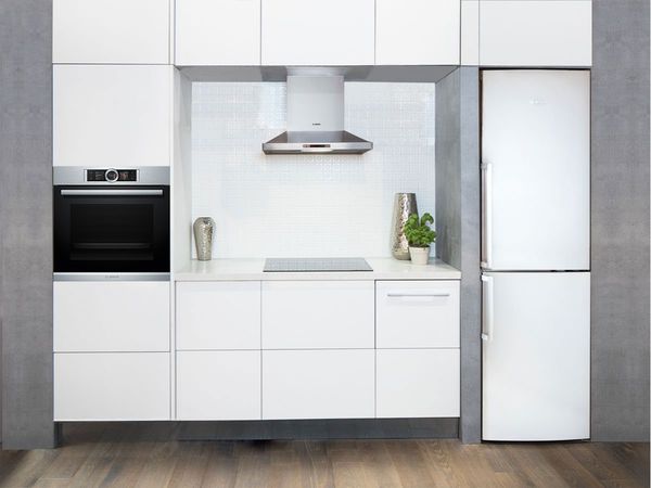 Minimālisma stila virtuve pilnīgi baltā krāsā, kuras centrā atrodas gatavošanas virsma un tvaika nosūcējs augšdaļā uz retro alvas krāsas fona
