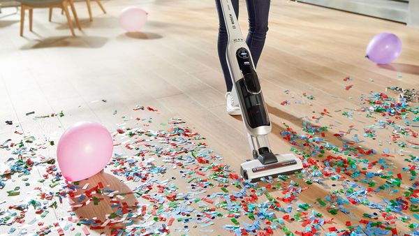 A woman vacuums confetti with a cordless vacuum cleaner. There are balloons everywhere.