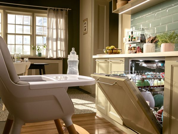 בקבוק לתינוקות ניצב על כיסא תינוק במטבח עם פינת אוכל. מדיח כלים של Bosch עם דלת פתוחה מעט, ולידו כיסא.