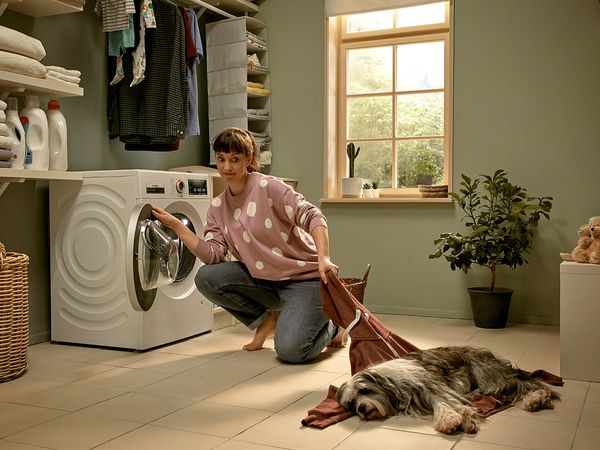Femei stând în fața unei mașini de spălat rufe, trăgând de un pulover pe care stă un câine.