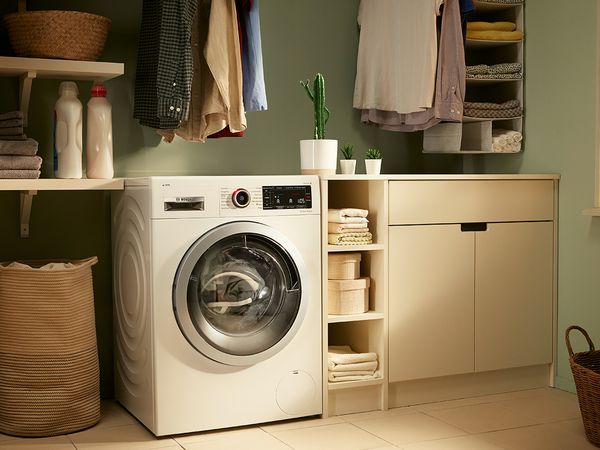 Una lavadora Bosch con dosificación inteligente i-DOS colocada en un cuarto de lavandería. Junto a la lavadora hay unos armarios y, encima de ellos, un cactus que parece hacer el gesto de la paz como si tuviera dos dedos.