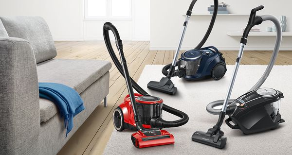 Three bagless vacuums in red, dark blue and black, forming a triangle on a beige living room carpet