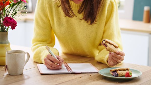 Eine Frau mit gelbem Pullover sitzt an einem Tisch mit Blumenstrauß, trinkt Tee und isst Gebäck von einem Teller. Sie schreibt mit einem Stift in ein Ringbuch.