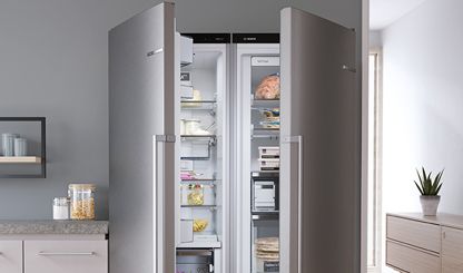 Серебряный отдельностоящий холодильник Bosch на белой кухне. Открытые дверцы и видно продукты. 