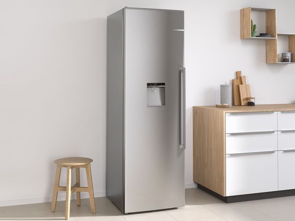 Brīvstāvošs sudraba krāsas Bosch ledusskapis starp nelielu tabureti kreisajā pusē un virtuves leti labajā pusē.