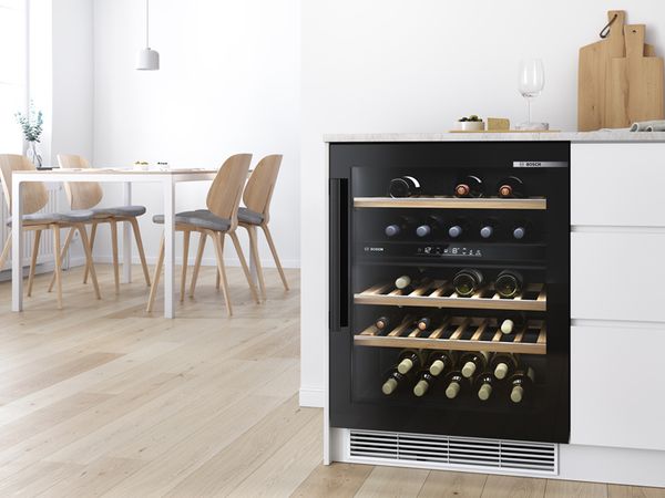 Шкаф Bosch для хранения и охлаждения вина со стеклянными дверцами демонстрирует коллекцию вина. Современный и светлый обеденный зал слева.