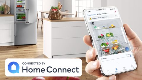 Bosch mosógép egy modern fürdőszobában, az előtérben egy kéz, benne eg okostelefon, rajta megnyitva a Home Connect mobilalkalmazás