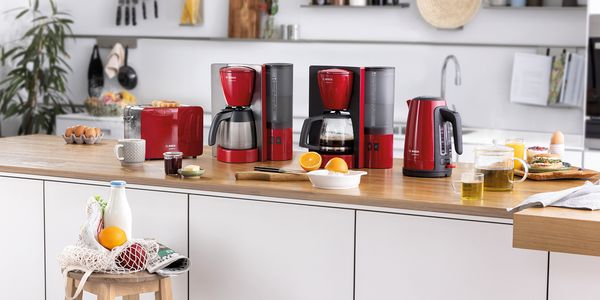 Komplet ComfortLine crvene boje i od nehrđajućeg čelika s tosterom, aparatom za filtar kavu i kuhalom za vodu. Na stolu su mnogi sastojci za doručak