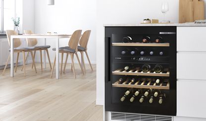 Шкаф Bosch для хранения и охлаждения вина со стеклянными дверцами демонстрирует коллекцию вина. Современный просторный и светлый обеденный зал слева.