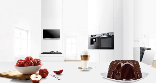 Un gâteau, des pommes et un café sur une table dans une cuisine blanche moderne avec plusieurs appareils électroménagers Bosch en arrière-plan.  