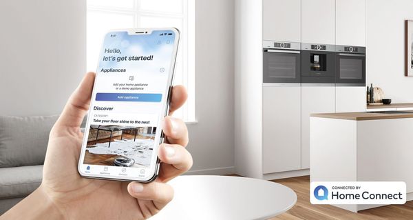 Dłoń trzymająca smartfona z zainstalowaną aplikacją Home Connect na tle nowoczesnej, białej kuchni.
