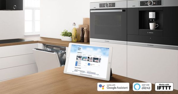 Tablet z otwartą aplikacją Home Connect na kuchennej wyspie; w tle urządzenia gospodarstwa domowego marki Bosch.