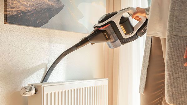 A woman is vacuuming a radiator with a cordless unlimited vacuum cleaner.