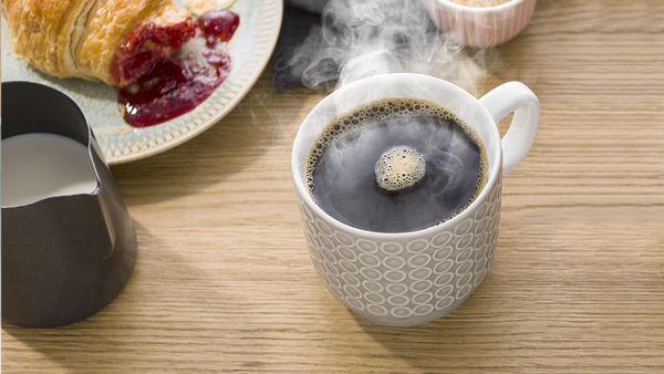Una humeante taza de café caliente, junto a un plato con un croissant y un tarro de leche.