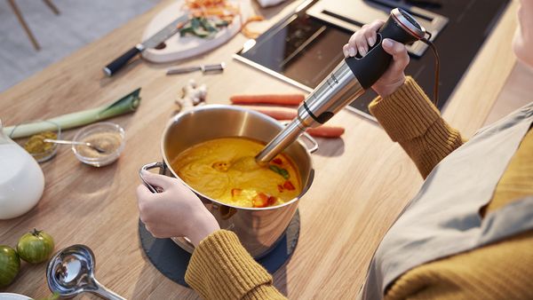 Une personne réduit en purée un pot de soupe de potiron avec un mixeur plongeant, avec des légumes frais autour.