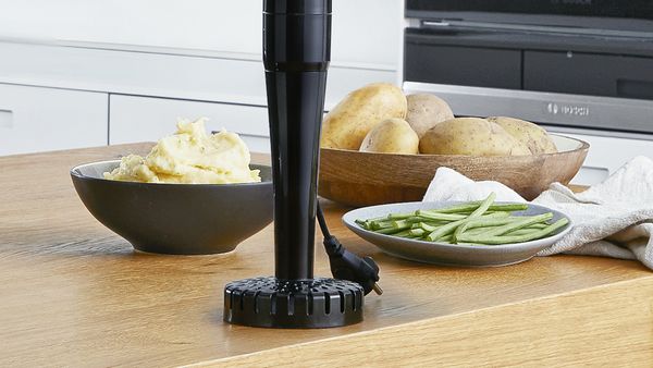A masher attachment for a hand blender on a table in front of various foods.