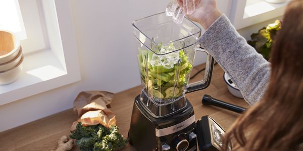 A person adding ice cubes to a Bosch blender to make a green smoothie.