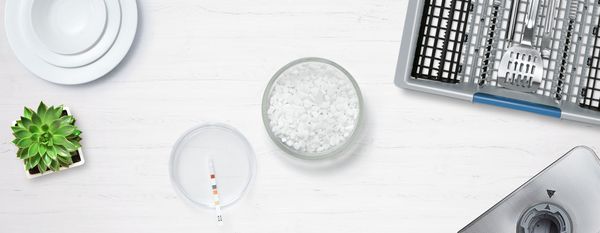 Sol za pomivalni stroj in testni listič za trdoto vode v posodah na belem pultu.