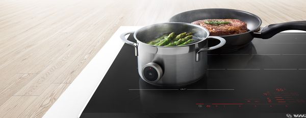 Индукционная варочная панель Bosch, кастрюля со спаржей и стейком на сковороде.