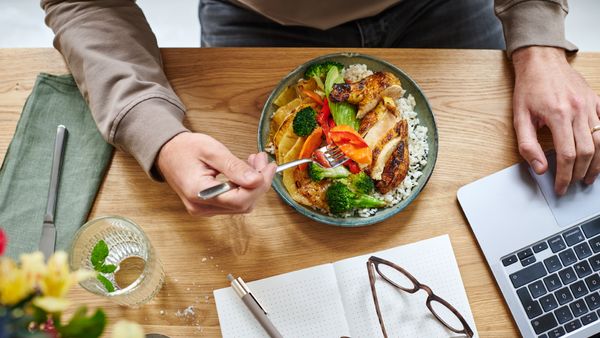 Eine Person sitzt am Schreibtisch vor einem aufgeklappten Laptop und isst aus einer Bowl ein Gericht mit Reis, Fleisch und Gemüse.