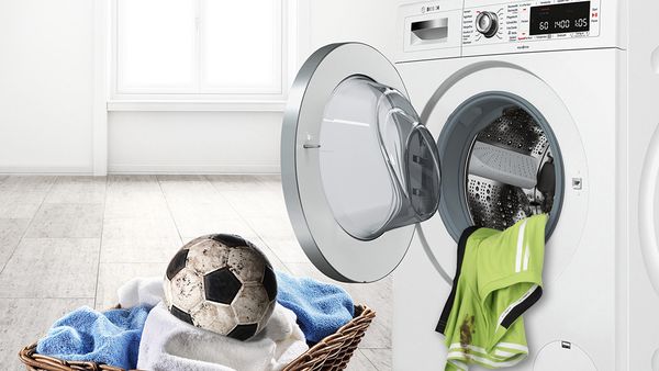 Špinavý fotbalový míč v koši na prádlo před pračkou s předním plněním.